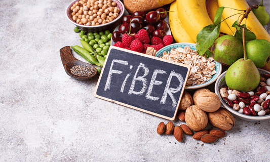 Alimentos ricos en fibra como frutas, legumbres, cereales integrales, frutos secos y semillas junto a cartel con la palabra fiber